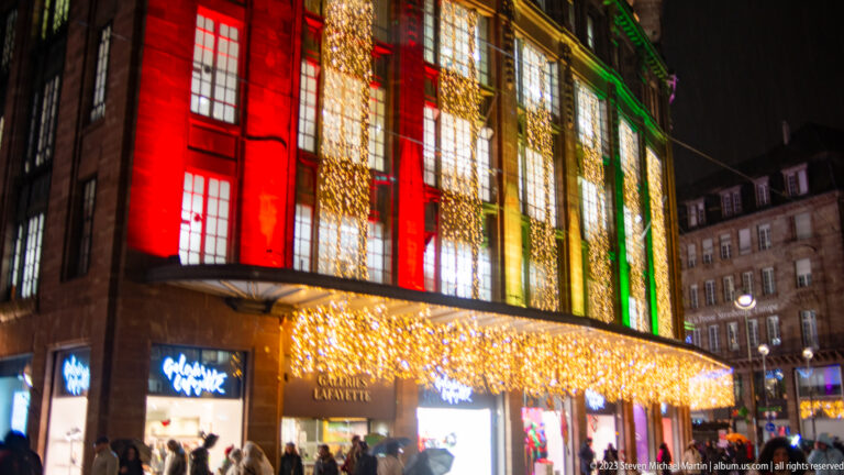 Galeries Lafayette Strasbourg façade lit in red, gold, and green with a glowing canopy over the entrance.