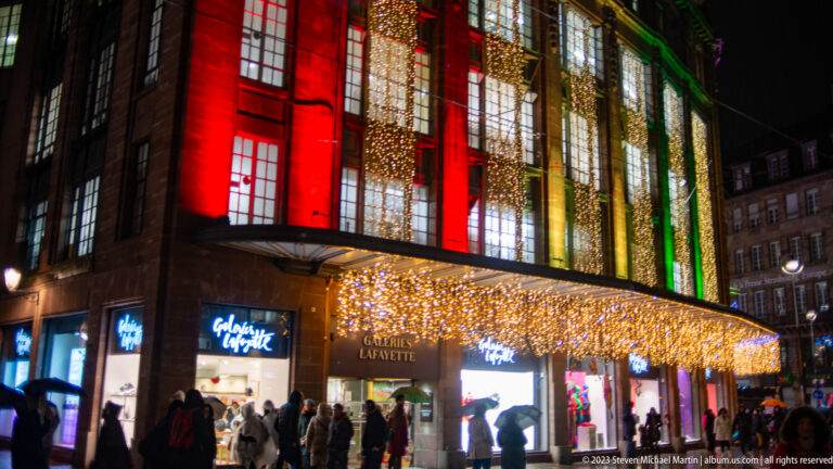 Wide view of Galeries Lafayette with shoppers beneath cascading warm lights and red, gold, and green façade illumination.