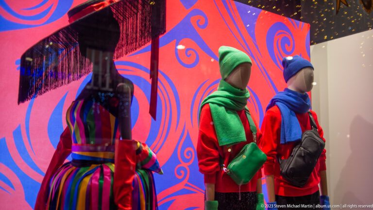 Two mannequins in red clothing with green and blue winter accessories beside a striped mannequin and vivid graphic backdrop.