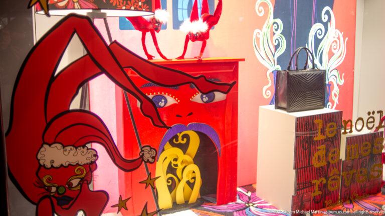 Colorful holiday shop window with red illustrated cabinet, whimsical figures, and stacked festive gift bags.