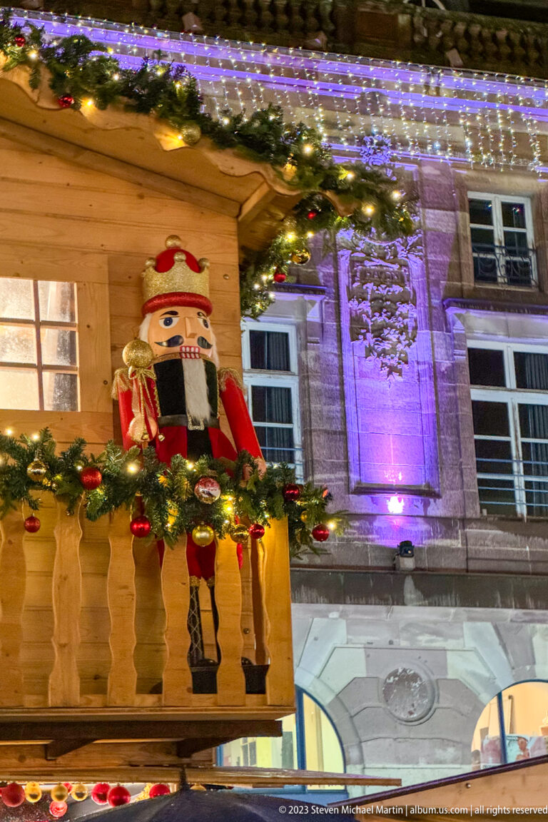 Large red-and-black nutcracker on a garland-decorated wooden balcony against a purple-lit stone façade.
