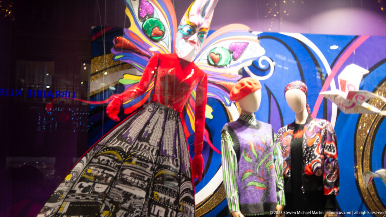 Two mannequins beside a tall masked theatrical figure in red and patterned clothing against a vivid blue backdrop.
