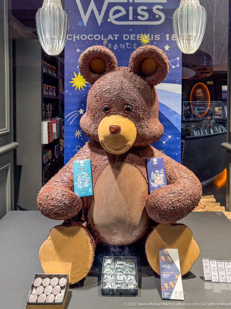 Large teddy bear in a Weiss chocolate shop window with boxed chocolates and confectionery displayed around it.