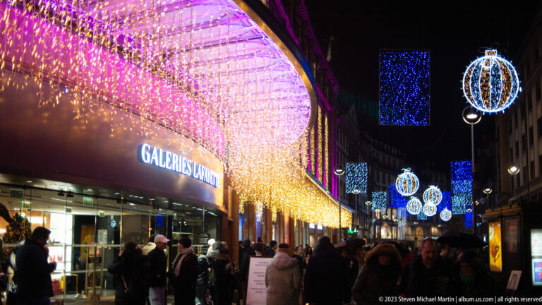 Galeries Lafayette entrance beneath pink, purple, and gold Christmas lights with blue hanging ornaments farther down the street.