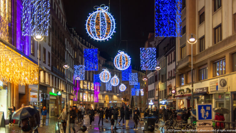 Wet Strasbourg pedestrian street with shoppers beneath blue light panels and hanging spherical Christmas ornaments.