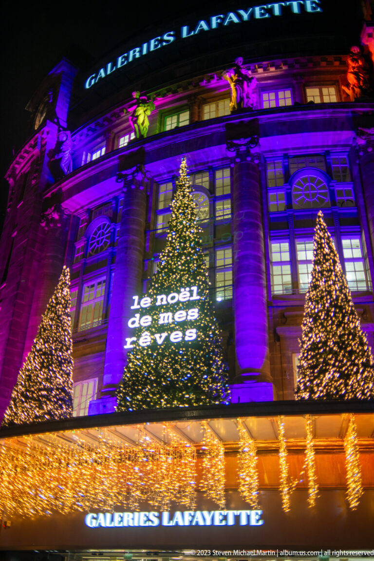 Galeries Lafayette façade with three Christmas trees, purple lighting, golden canopy, and projected “Le Noël de mes rêves” message.