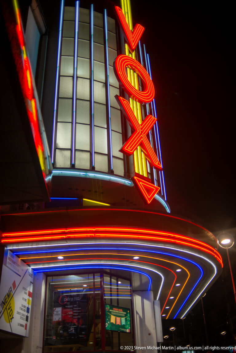 Vox cinema entrance with vertical red neon sign and curved canopy outlined in red, blue, and yellow lights.