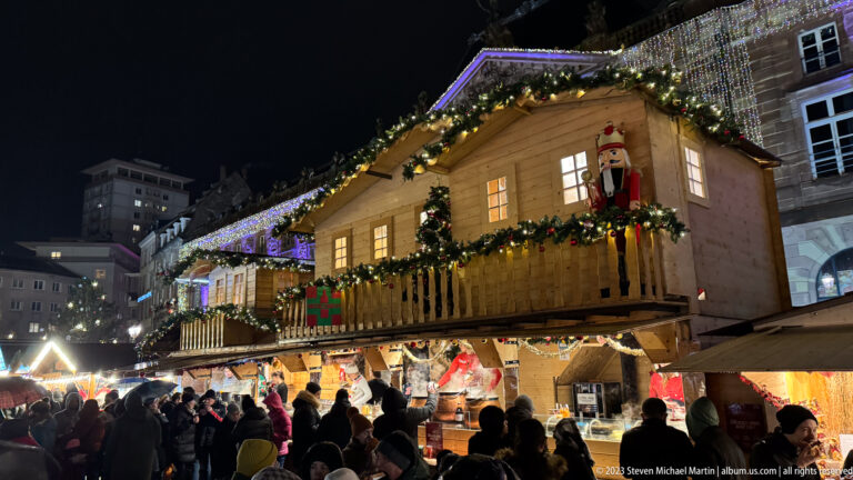 Two-story wooden Christmas chalet with balcony garlands, nutcracker figure, bright counters, and a dense crowd.