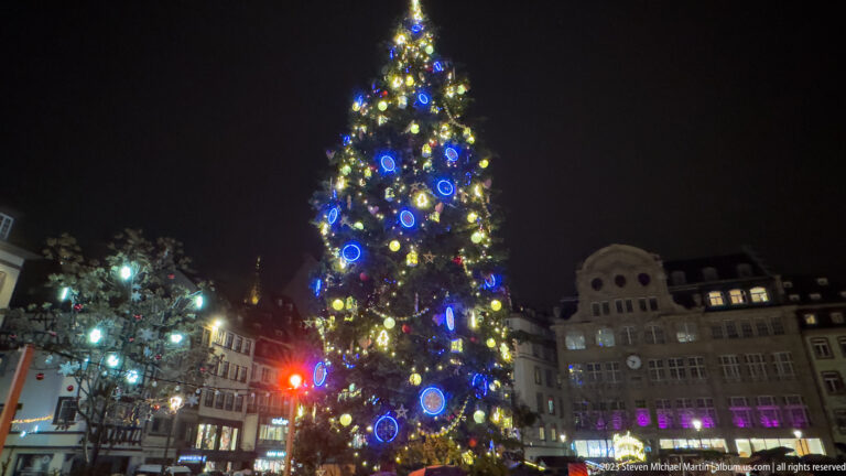 Tall illuminated Christmas tree with blue circular ornaments rising above Place Kléber at night.