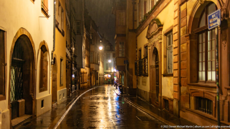 Narrow rain-slicked Strasbourg street lined with historic stone façades, arched openings, and warm lamps at night.