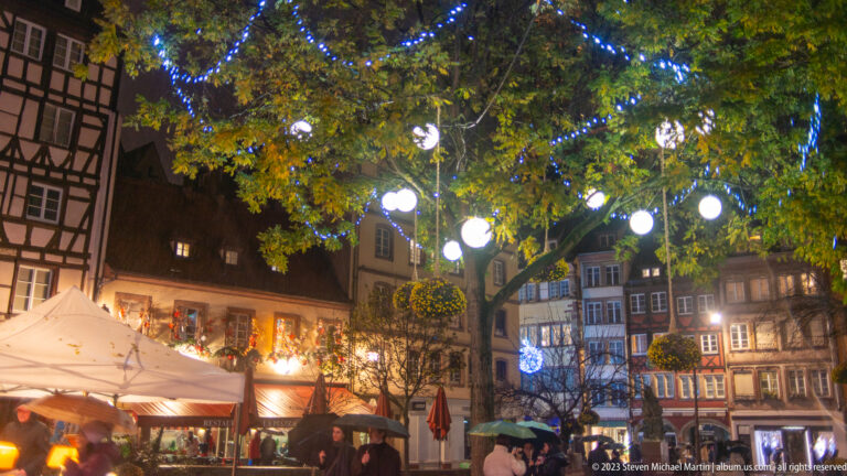Rainy Christmas-market square with umbrellas, half-timbered façades, warm stalls, and blue-and-white lights in tree branches.