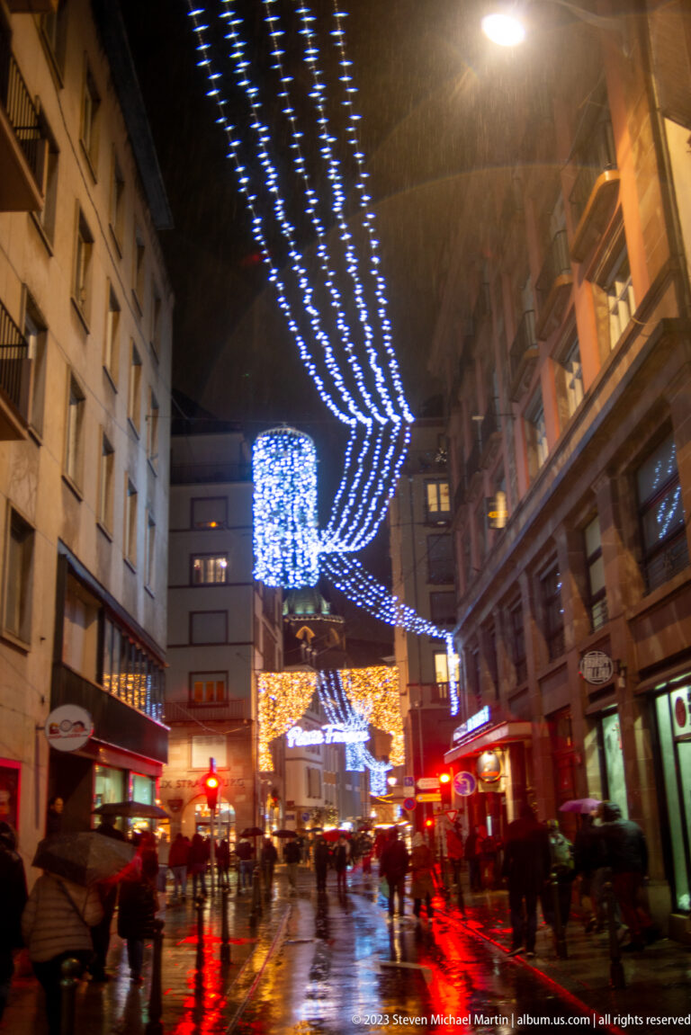 Wet Strasbourg street with pedestrians beneath blue light curtains and large suspended Christmas decorations.