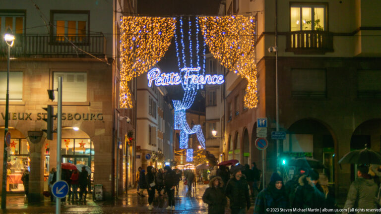 Illuminated “Petite France” sign above a narrow wet street with pedestrians, umbrellas, and blue-and-gold Christmas lights.