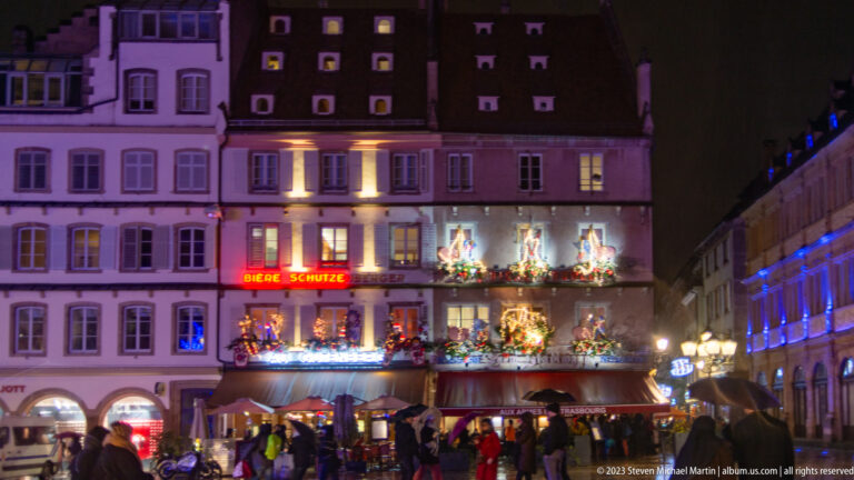 Historic façade covered with garlands, wreaths, figurative Christmas decorations, purple light, and restaurant signage.