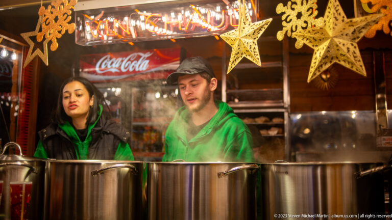 Two vendors behind steaming metal pots in a Christmas-market stall decorated with hanging stars.