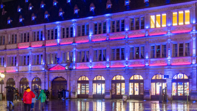 Long historic Strasbourg façade illuminated blue and pink, reflected on wet pavement with pedestrians in front.