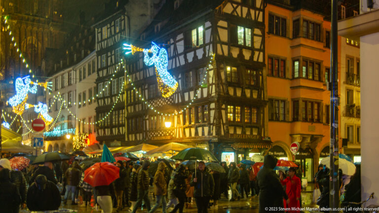 Crowded rainy Petite France street with half-timbered buildings and large blue angel-shaped Christmas lights overhead.