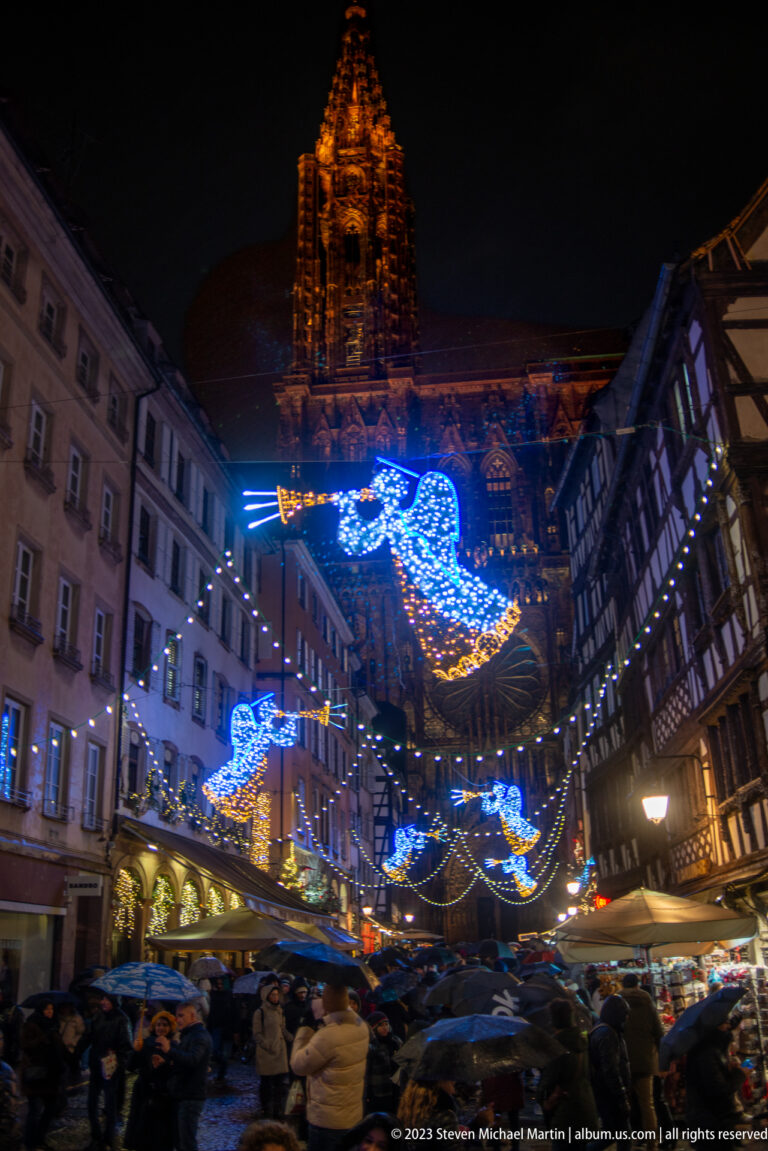 Strasbourg Cathedral tower above a crowded street lined with half-timbered buildings and blue-and-gold angel lights.