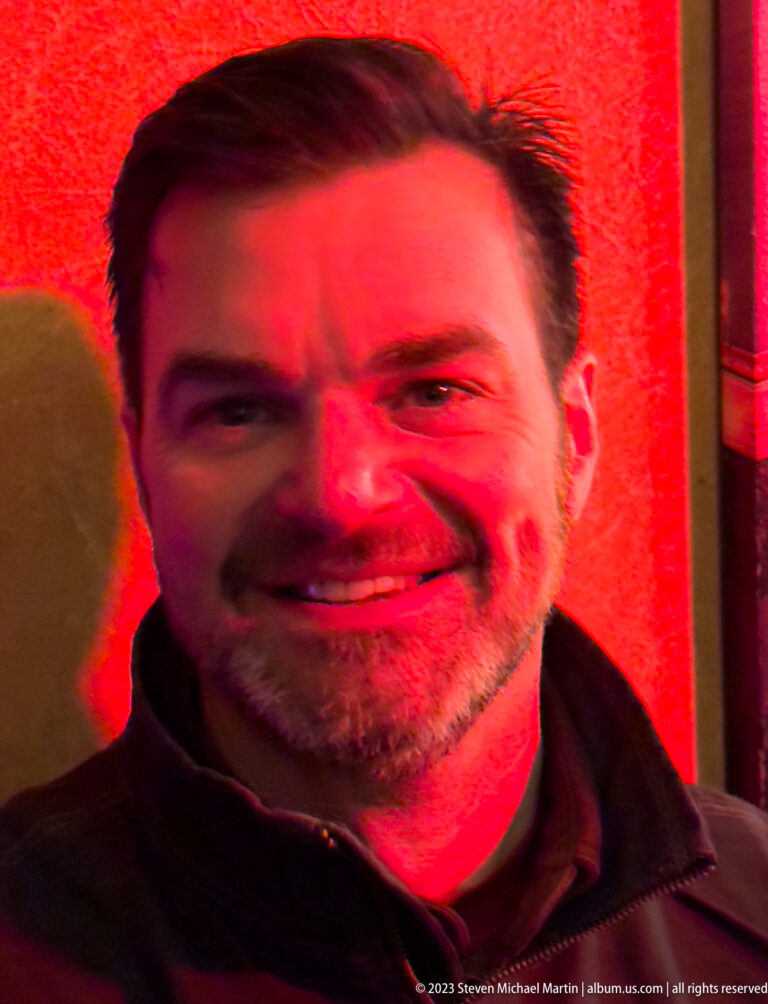 Close portrait of a smiling man with short dark hair and beard illuminated by strong red light.