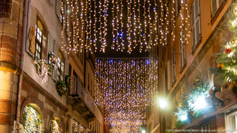 Narrow Strasbourg lane beneath dense warm-white hanging light curtains, with wreaths and decorated windows along the façades.
