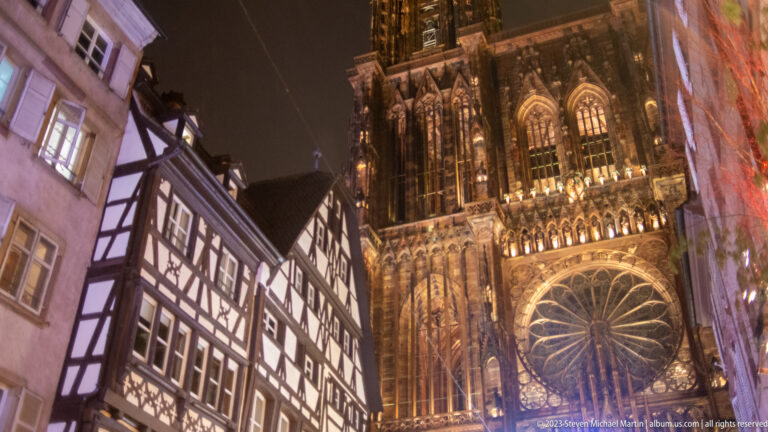 Strasbourg Cathedral rose window and Gothic west façade rising beside a half-timbered house at night.