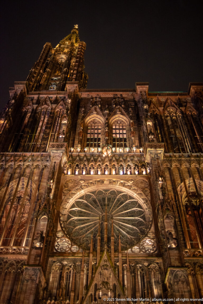 Low-angle night view of Strasbourg Cathedral’s rose window, tracery, pinnacles, and illuminated upper west façade.