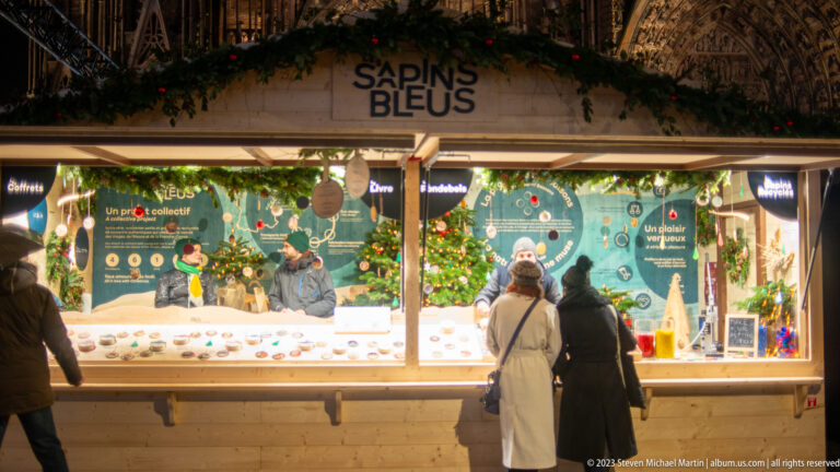 Visitors at a Christmas-market chalet labeled “Sapins Bleus,” with green illustrated panels and evergreen garland.