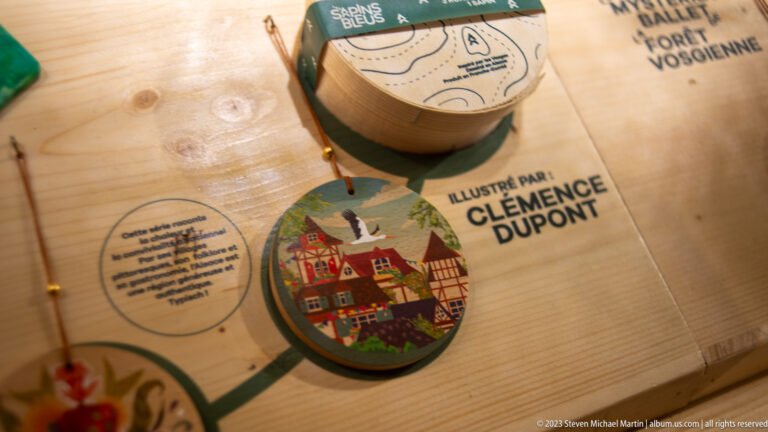 Round wooden ornament with colorful Alsatian buildings beside printed credit “Illustré par Clémence Dupont.”