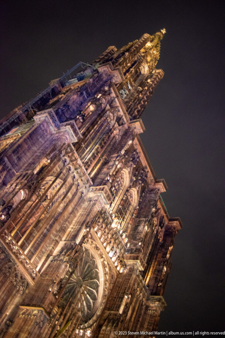 Oblique low-angle night view of Strasbourg Cathedral with rose window, Gothic tracery, buttressing, and tower.