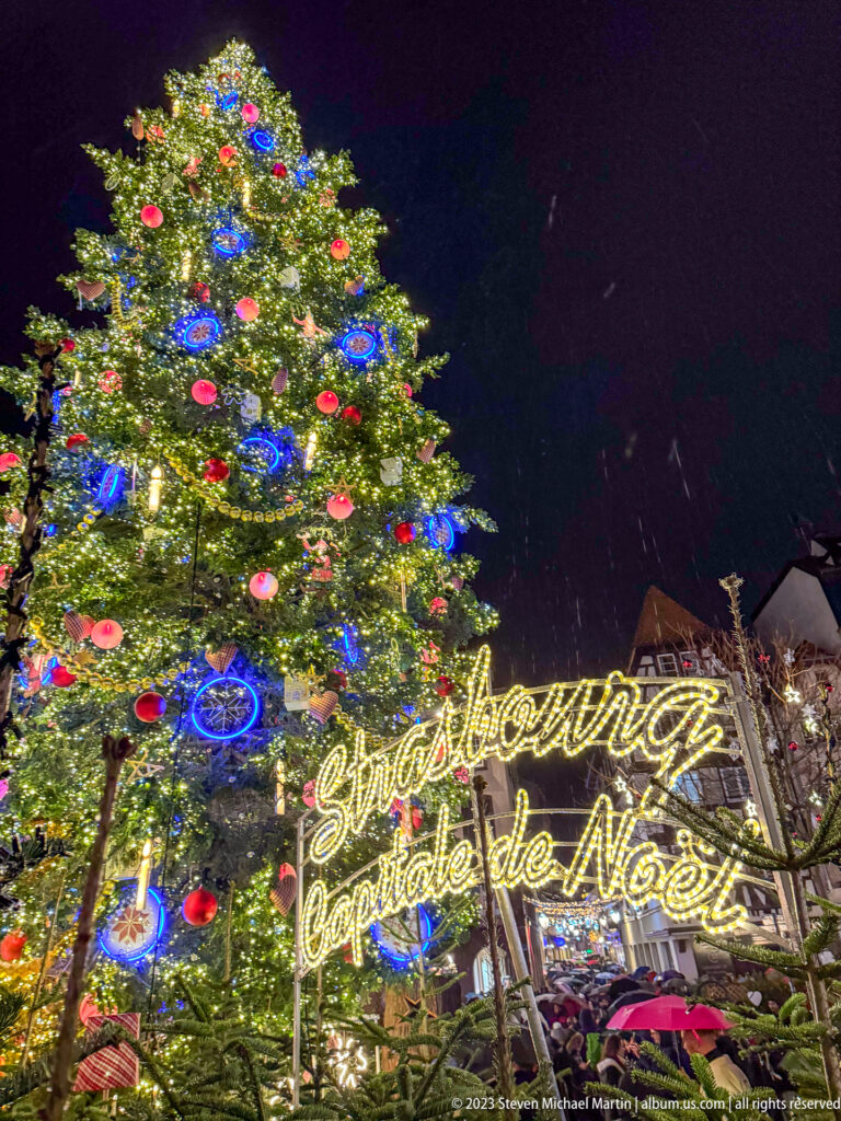 Illuminated Christmas tree beside glowing script reading “Strasbourg Capitale de Noël” at night.