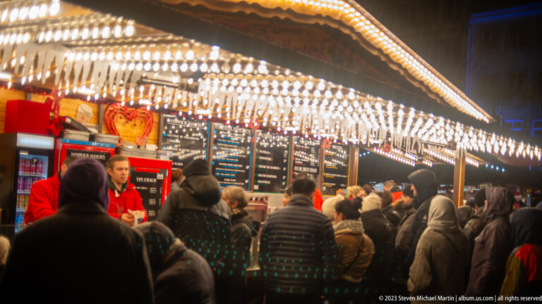 Crowded Christmas-market counter with bright bulb canopy, menu boards, customers, and staff behind the bar.