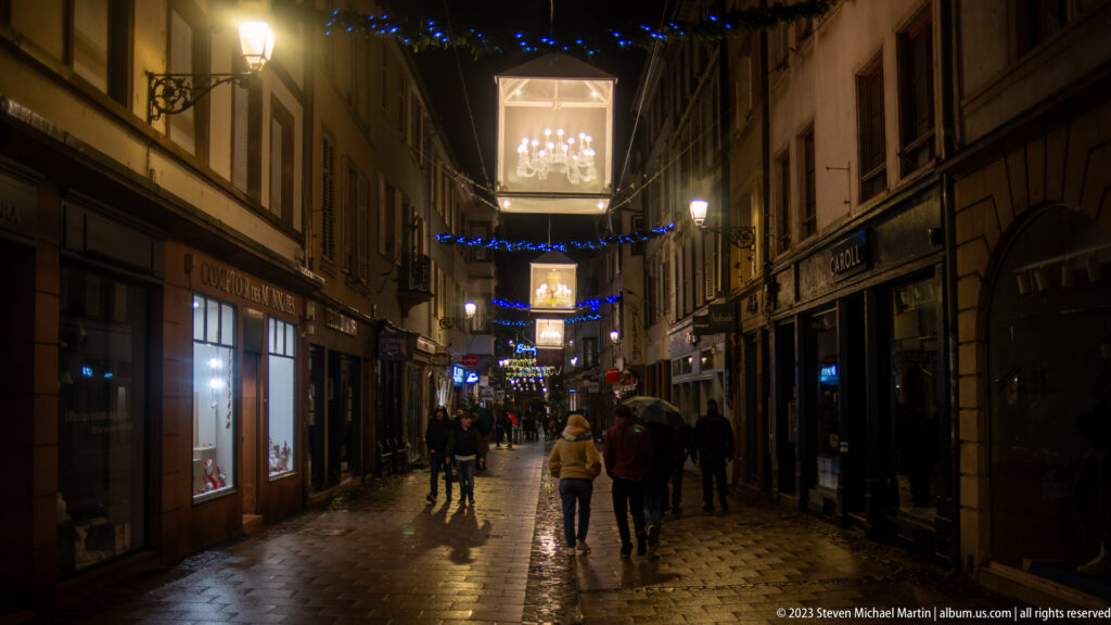 Narrow wet Strasbourg pedestrian lane with suspended illuminated cubes, blue garlands, shops, and walkers.