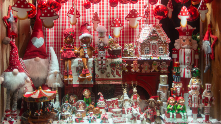 Crowded Christmas display with gingerbread house, gnomes, nutcrackers, mushrooms, and miniature figures against red gingham.