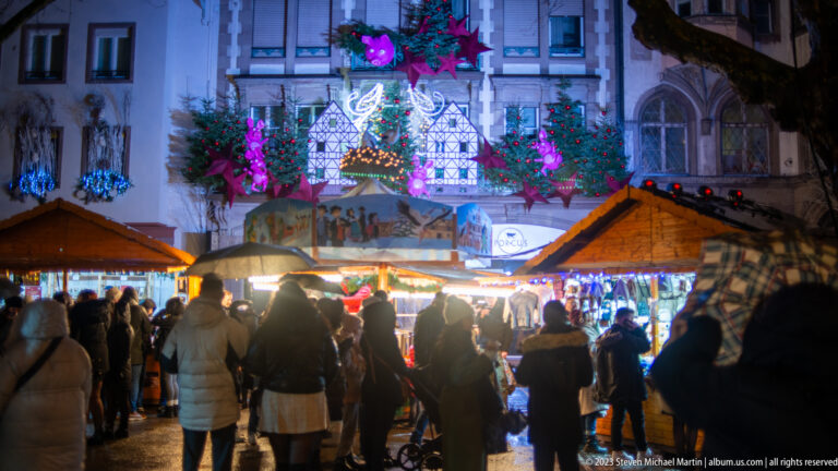 Crowded Strasbourg Christmas market with wooden chalets beneath a large colorful illuminated façade decoration.
