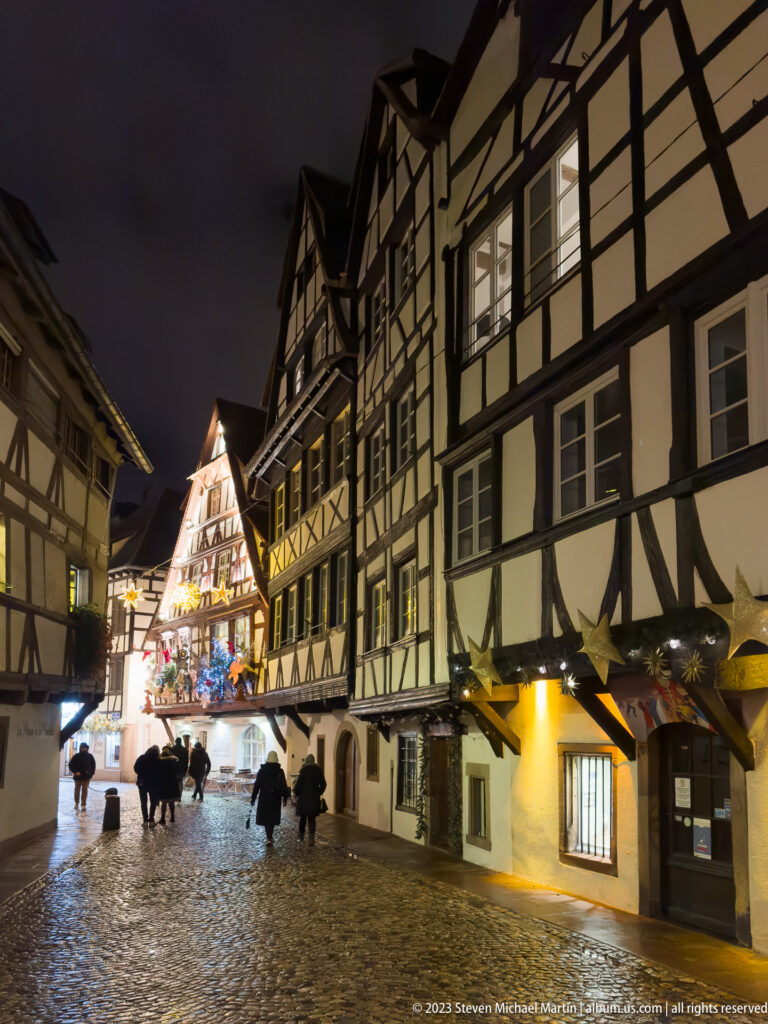 Wet cobbled Strasbourg square lined with half-timbered houses, umbrellas, and a brightly illuminated gabled building ahead.