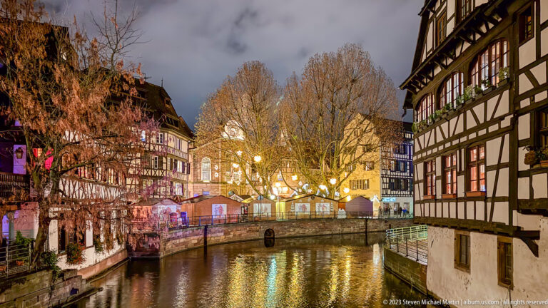 Petite France canal at night with half-timbered houses, leafless trees, market huts, and colorful reflections.