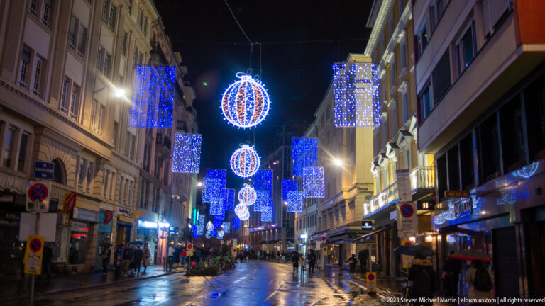 Wet Strasbourg street beneath blue rectangular Christmas lights and hanging glowing spheres at night.