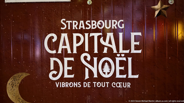 Red sign reading “Strasbourg Capitale de Noël — Vibrons de tout cœur,” with star and crescent decorations.