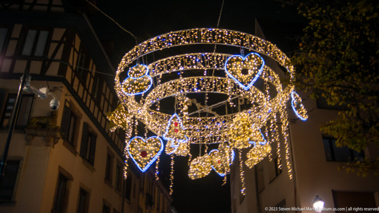 Circular overhead Christmas light installation with glowing hearts, stars, and hanging ornaments above a narrow street.