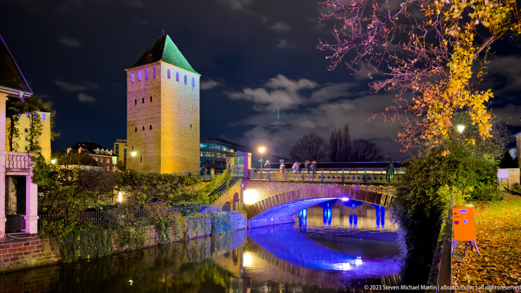 Square medieval tower with green roof beside a bridge and canal, with blue light reflected in the water.
