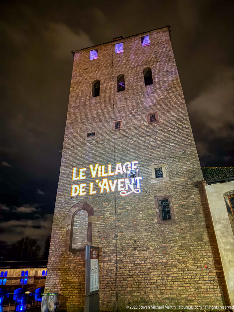 Tall masonry tower with “Le Village de l’Avent” projected across its wall and purple-lit upper windows.