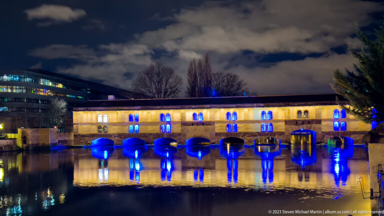 Barrage Vauban at night with warm-lit façade, blue arches, and strong reflections in the River Ill.