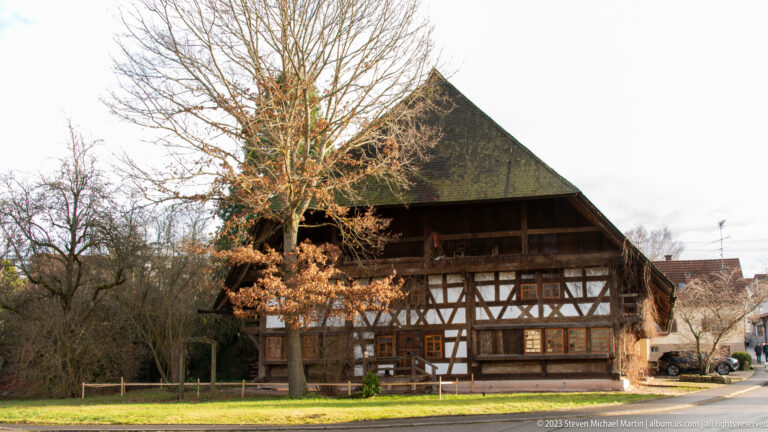 Traditional Black Forest farmhouse with half-timbered walls, broad dark roof, wooden gallery, and leafless trees.