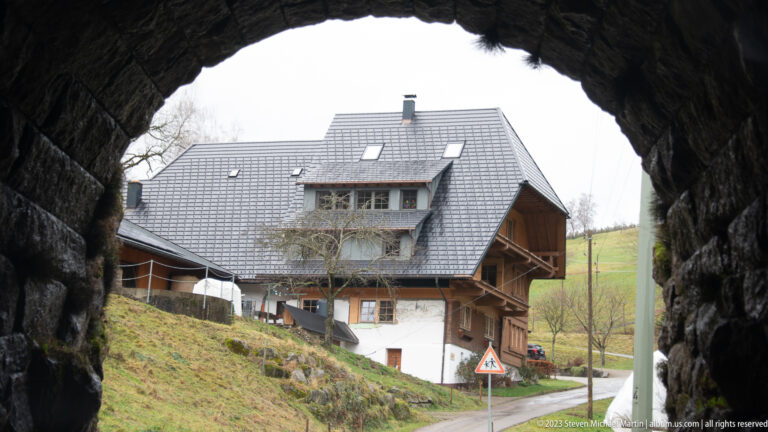 Traditional steep-roofed house with timber balconies viewed through a dark rough-stone archway.