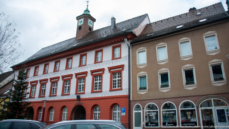 Red-and-white historic town building with arched ground floor, tall windows, tiled roof, and small clock turret.