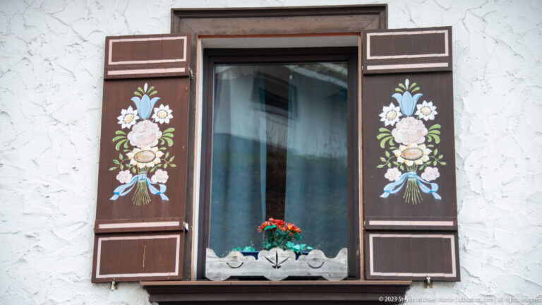 Window with dark brown shutters painted with colorful flower bouquets on a textured white stucco wall.