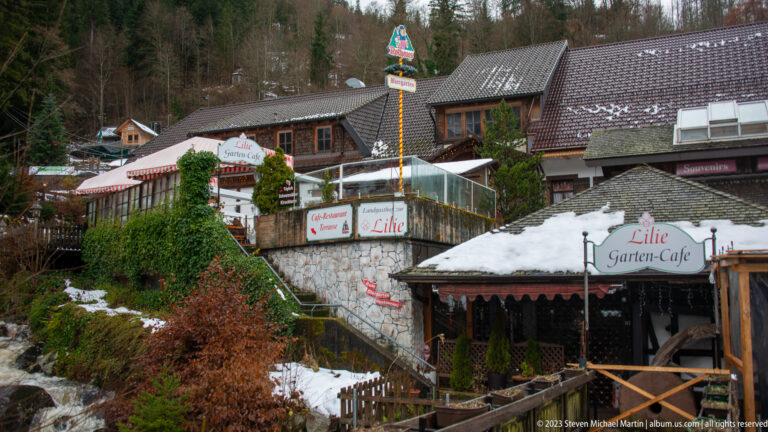 Terraced Lilie Garten-Café in Triberg with rustic buildings, stone walls, outdoor structures, and patches of snow.