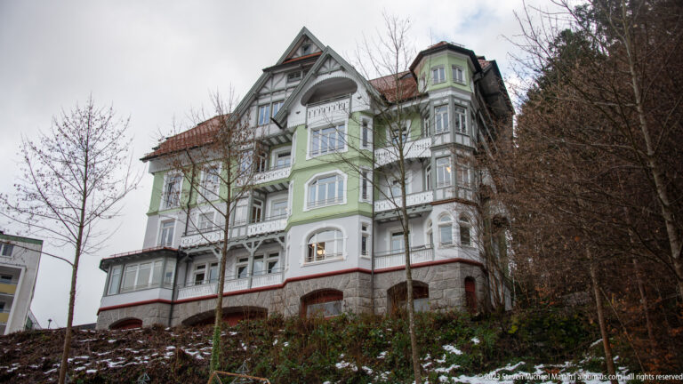 Tall pale-green hillside villa with white timber ornament, projecting bays, balconies, and steep gables among bare trees.