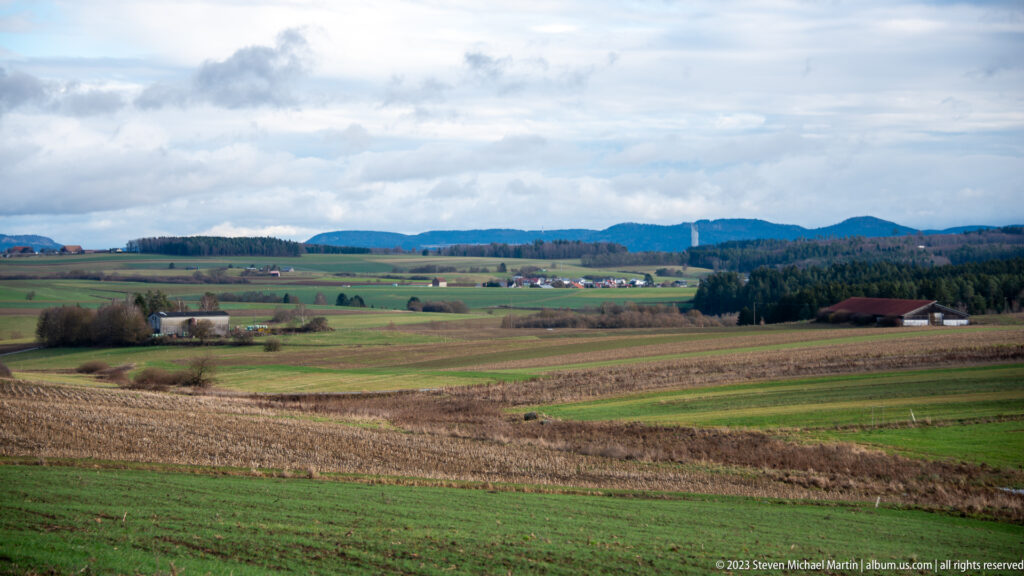 Rolling green and brown farm fields with scattered buildings below dark forested ridges under a cloudy sky.