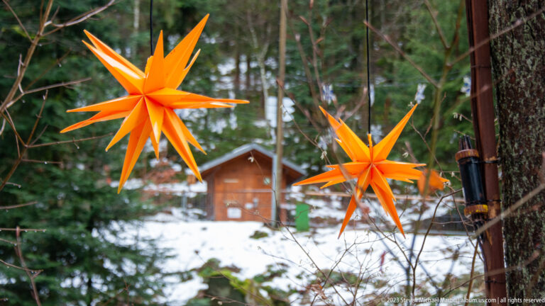 Two bright orange many-pointed Christmas stars hanging among trees, with snow and a small wooden cabin behind.